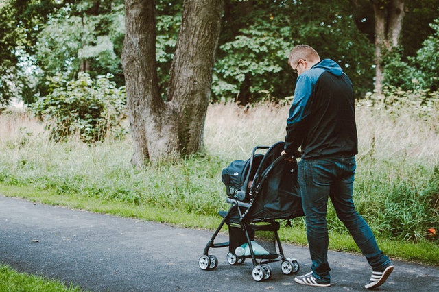 A father pushing his baby in a pram through a park surrounded by tall green grass and trees