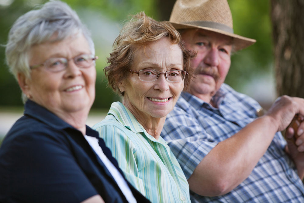 Two women and a man are sitting on a park bench, smiling at the camera.