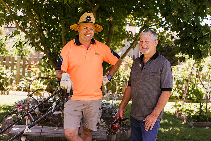 A senior man smiles at the camera. He is standing next to an LCHS staff member wearing an orange high vis shirt. The staff member is leaning against a lawn mower and wearing gardening gloves