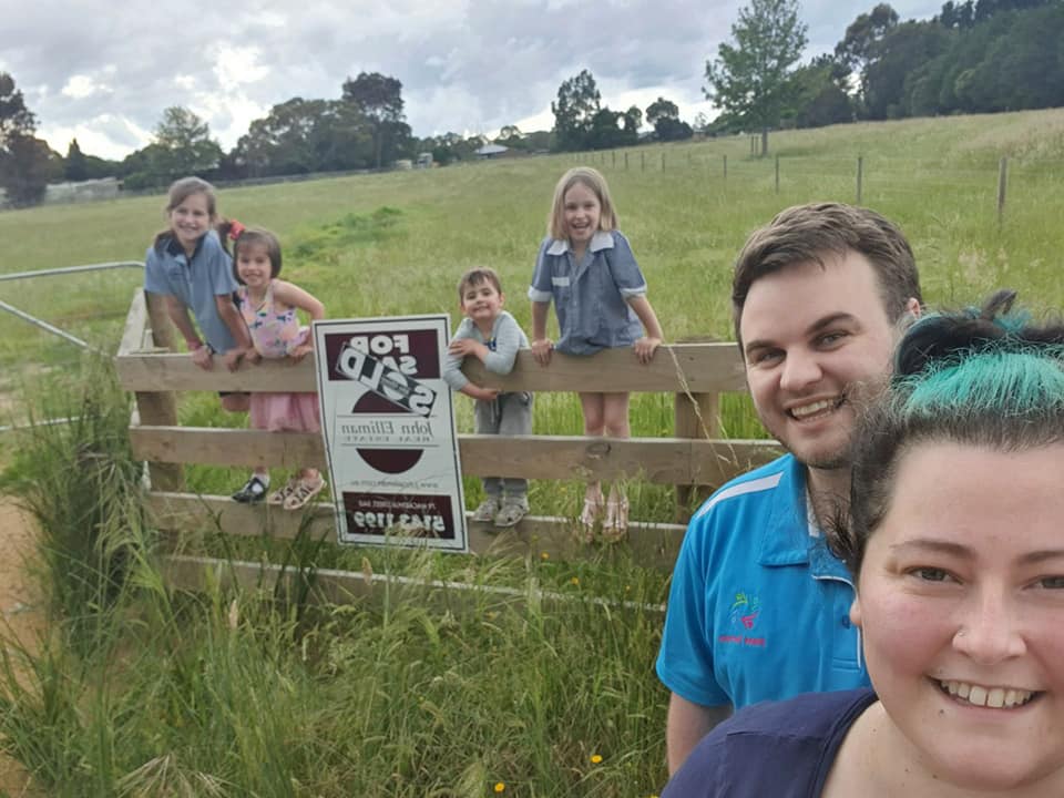 Pictured is Jamie and his wife standing in front of a sold sign on a block of land. Their four children are climbing the fence around the sold sign. Everyone is smiling at the camera.