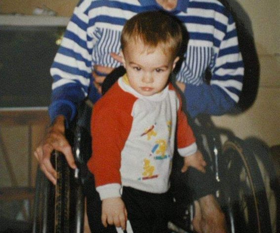 Pictured is 3 year old Jaime standing in front of a man in a wheelchair. Jamie is staring directly at the camera looking apprehensive.