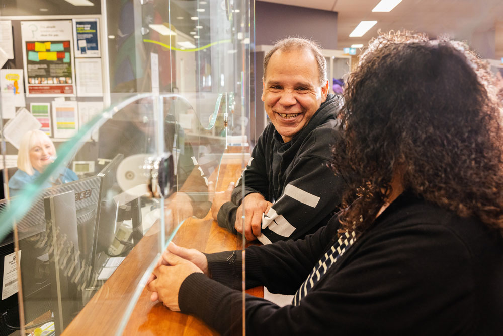A First Nations man waits at the reception counter with a woman, smiling. A First Nations man waits at the reception counter with a woman, smiling.