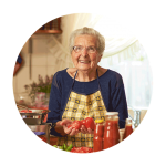 A senior women wearing glasses and and apron holds chopped tomatoes while smiling at the camera