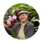A senior man sits in a garden and holds a pink blooming orchid. He is wearing a hat and glasses and smiles at the camera